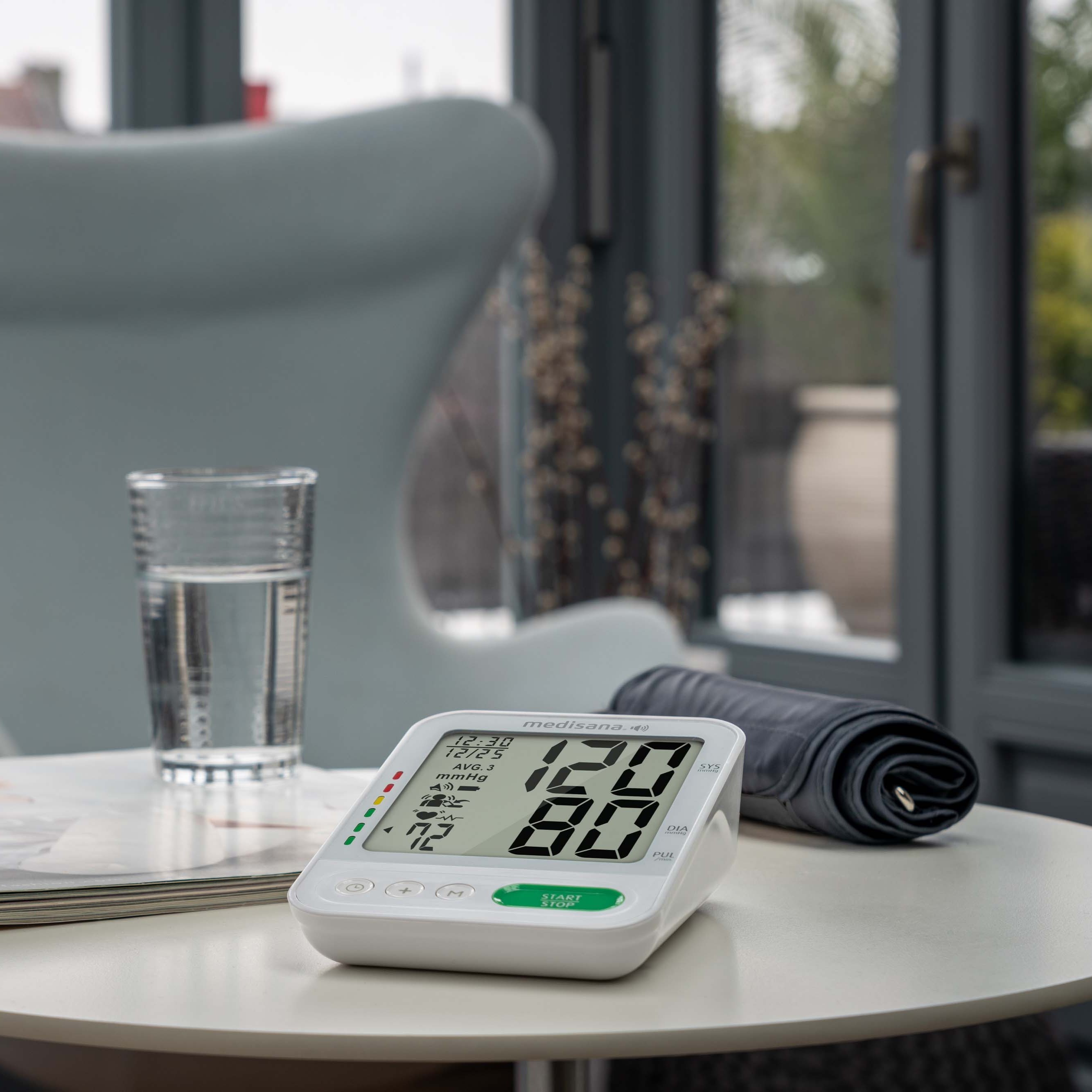 A digital blood pressure monitor sits ready for use on a small table, with the cuff placed beside it and a glass of water nearby.